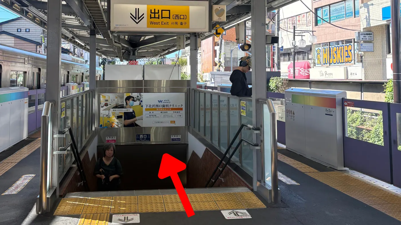 京王井の頭線のホームで「出口（西口）」の案内板に従って階段を下りる様子（矢印は進行方向）