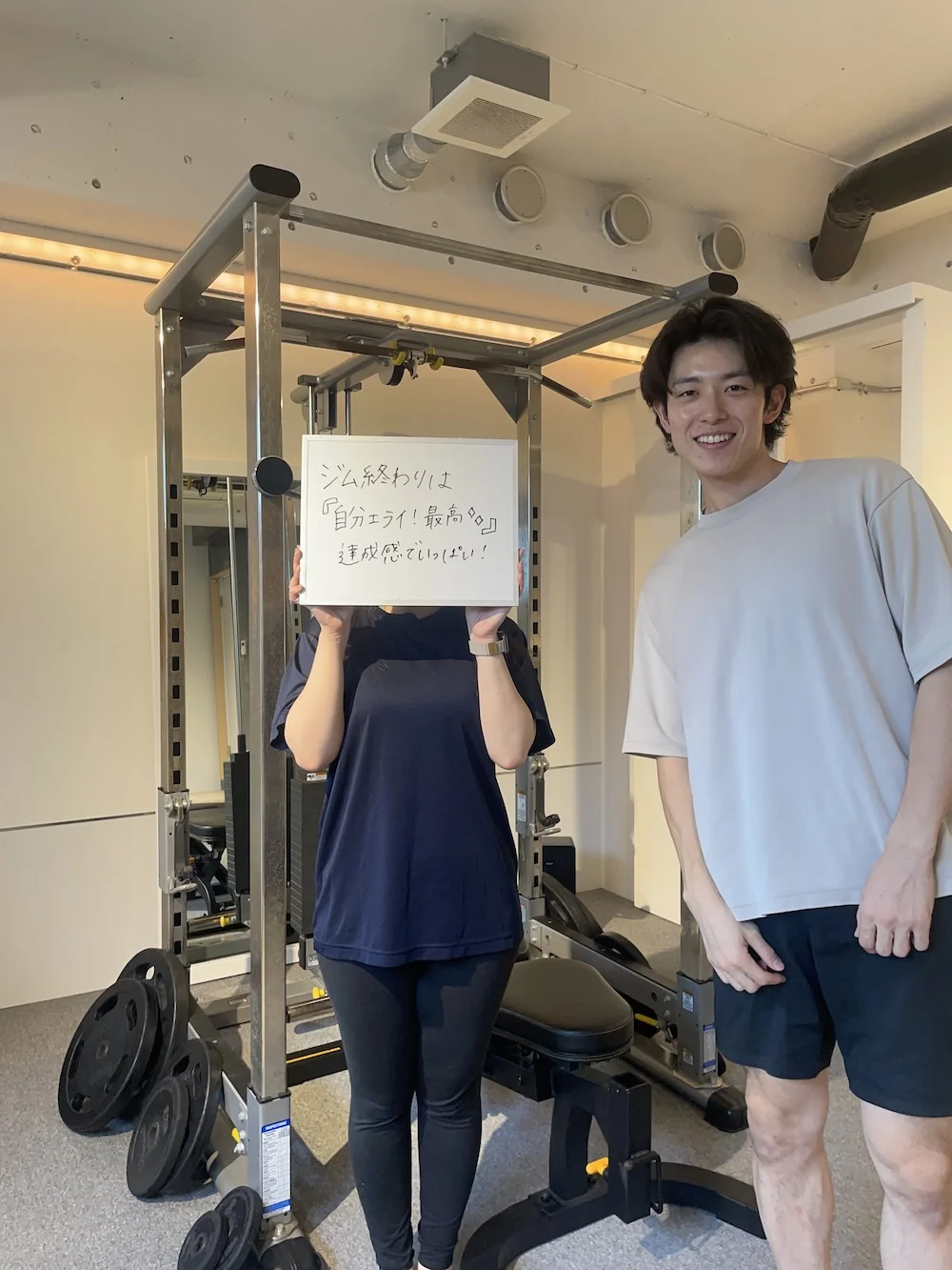 WORKOUTHOLIC下北沢店で「ジム終わりは自分エライ！最高◎達成感でいっぱい！」と書いたホワイトボードを掲げる20代女性会員と、隣で笑顔のトレーナーの写真