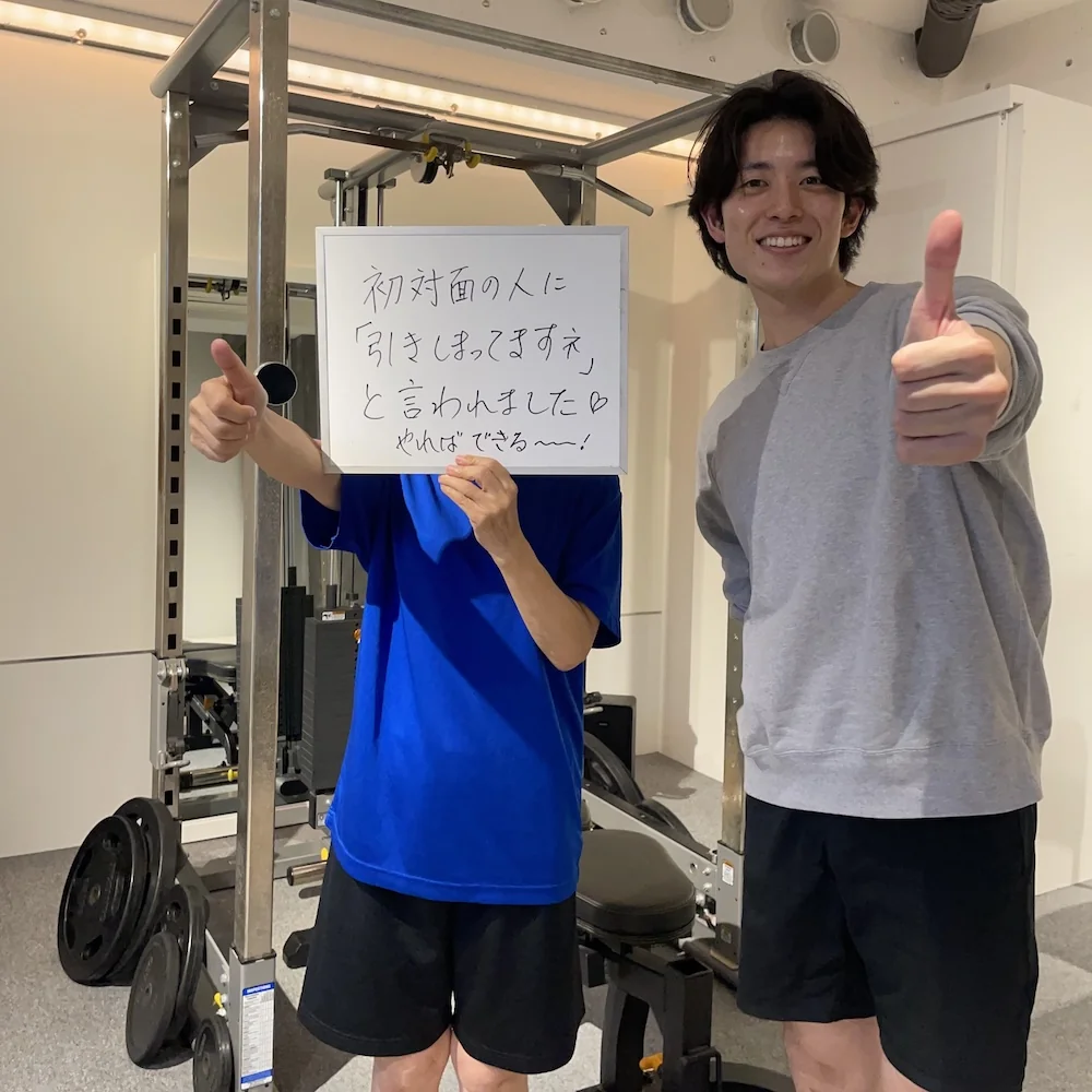 WORKOUTHOLIC下北沢店で「初対面の人に引きしまってますねと言われました、やればできる〜〜！」と書いたホワイトボードを掲げる50代女性会員と、隣でサムズアップするトレーナーの写真
