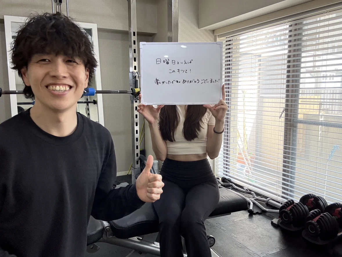 WORKOUTHOLICのトレーニングルームで、トレーナーと一緒に親指を立てる20代女性会員。「日曜日といえばこのキツさ！楽しかったです。ありがとうございました！」と書いたホワイトボードを掲げている