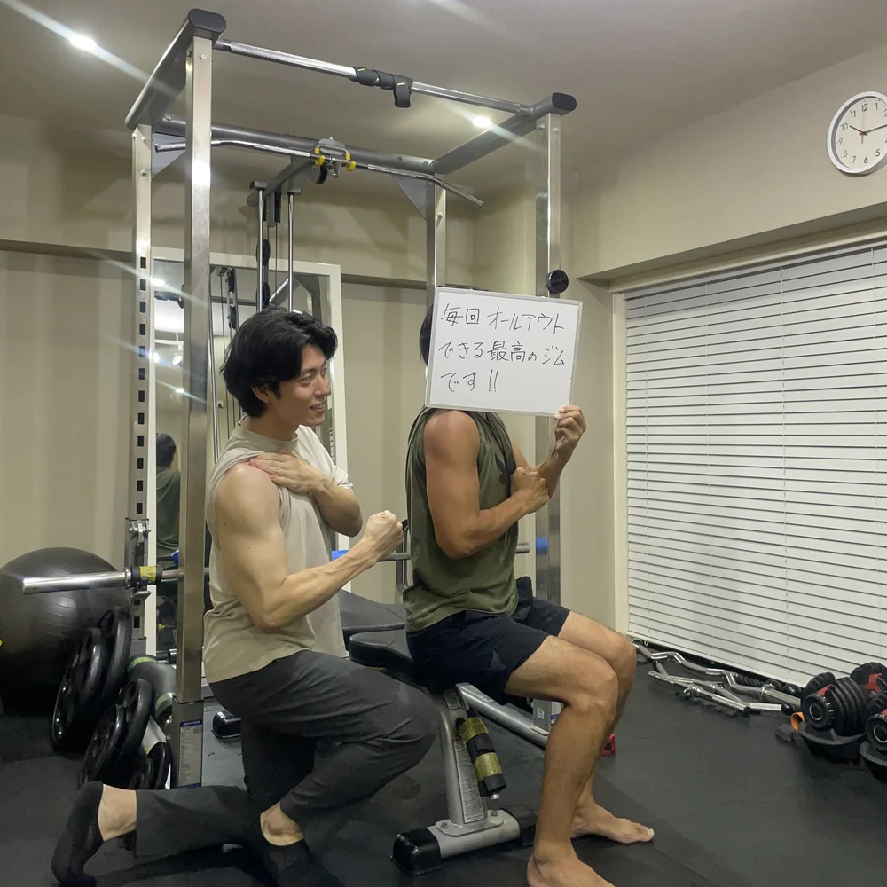 WORKOUTHOLICのトレーニングルームで、トレーナーと一緒にトレーニングする40代男性会員。「毎回オールアウトできる最高のジムです!!」と書いたホワイトボードを掲げている